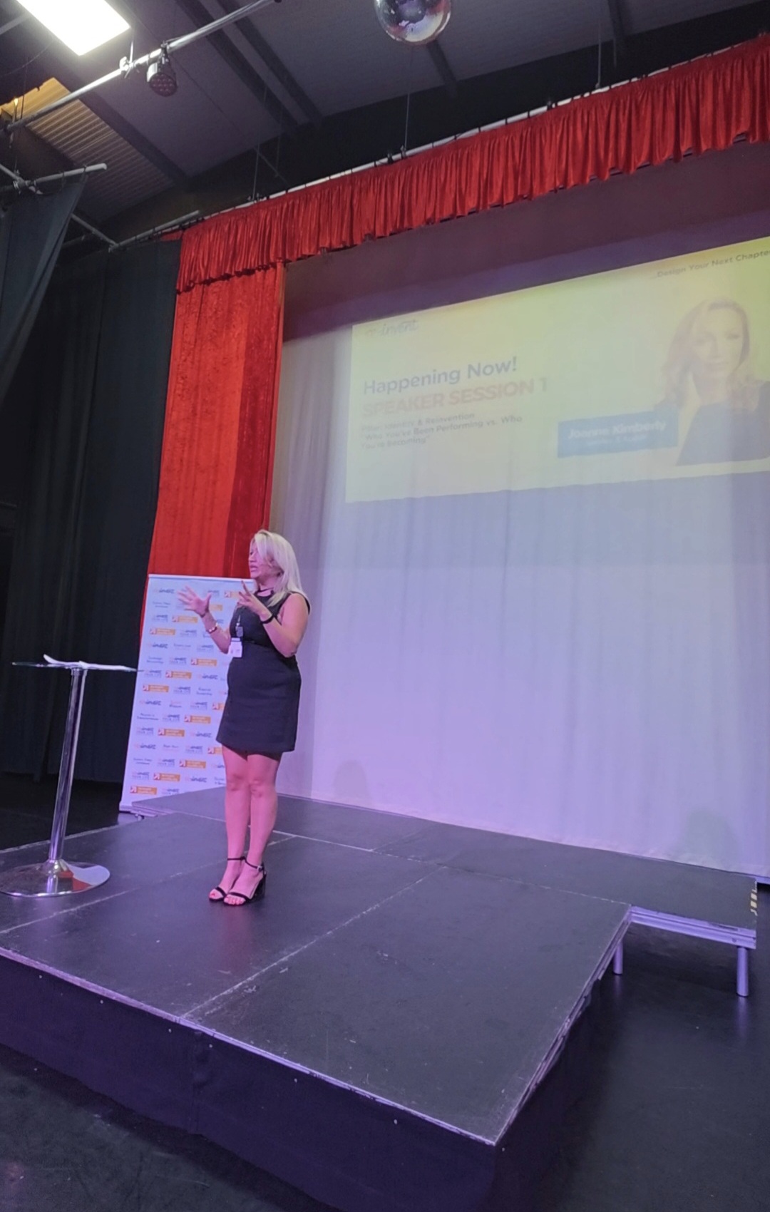 Joanne Kimberley speaking on a stage with a screen reading Speaker Session 1 behind her