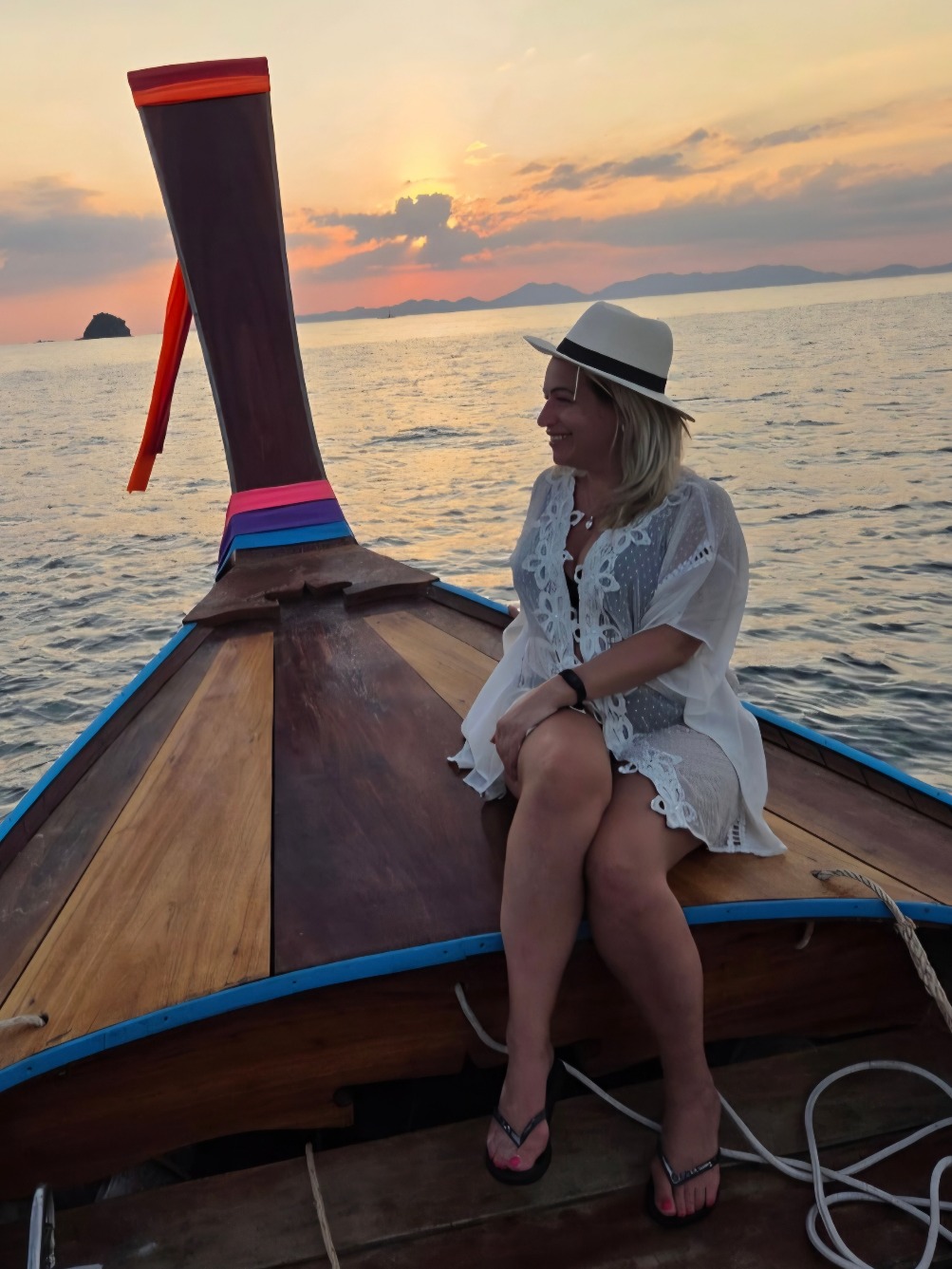 Joanne Kimberley in a white cover-up and sun hat seated on the bow of a traditional long-tail boat at sunset